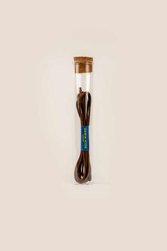 Elegant brown waxed cotton shoe laces polished with beeswax, displayed inside a stylish Oswin Hyde glass tube.