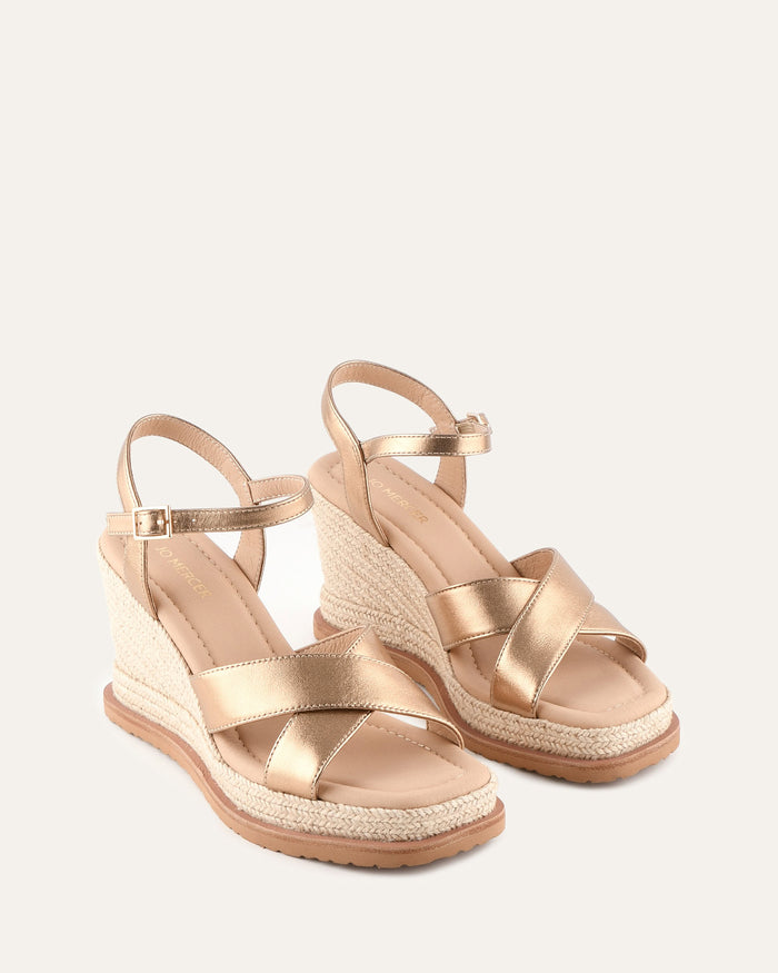 Pair of gold wedge sandals with espadrille sole on a light beige background