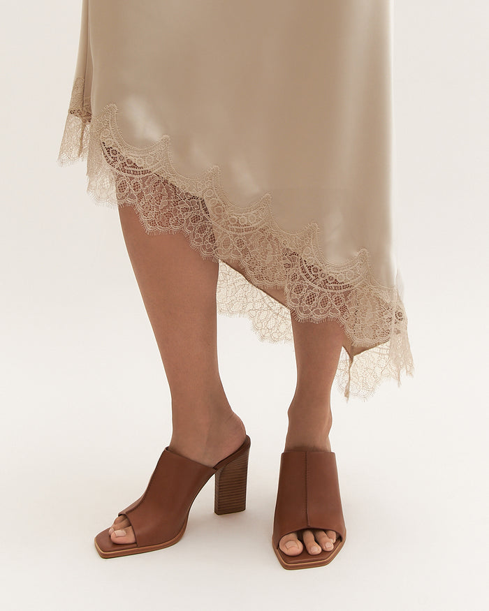 Person wearing a beige lace-trimmed dress with brown open-toe heels on a white background